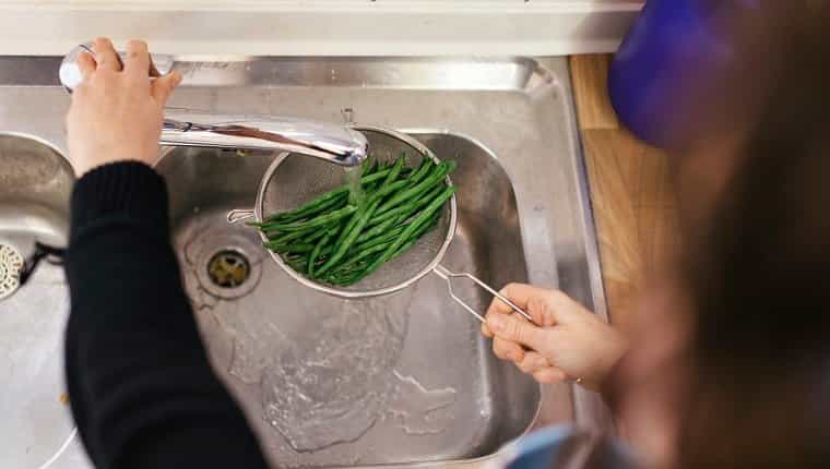 Cerca de judías verdes que se lavan con agua en el fregadero de la cocina.  Los mantiene bajo el agua una mujer que no se ve en la foto.