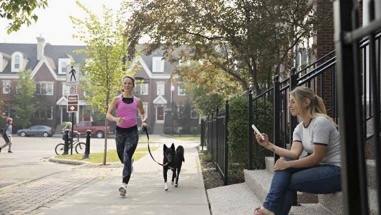 Corredoras corriendo con un perro y una mujer enviando mensajes de texto con su teléfono celular en la acera del barrio